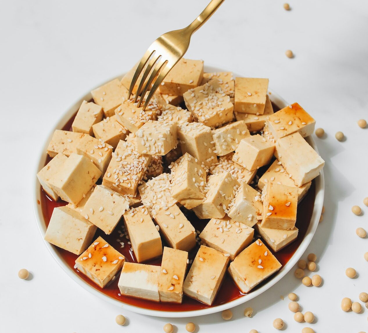 Person Holding Golden Fork and Brown Tofu on White Plate
